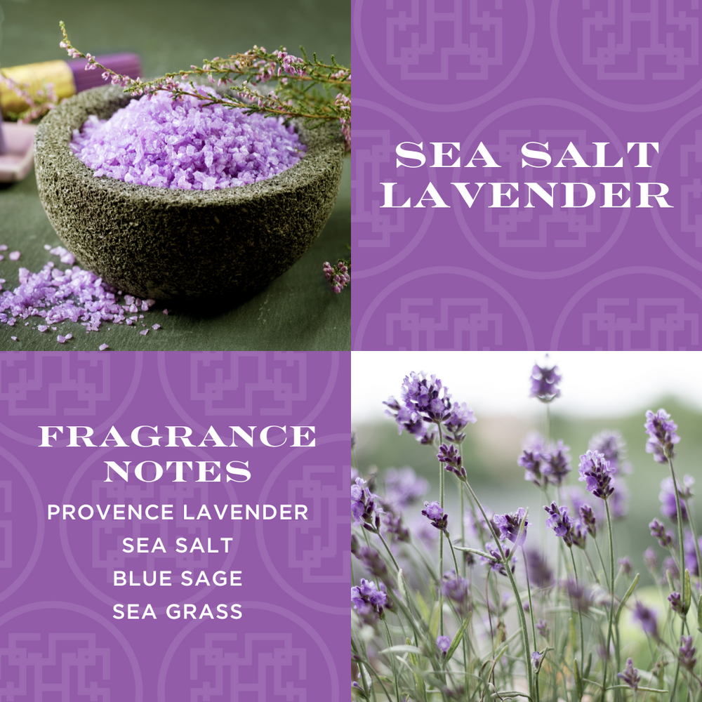 A candle with lavender-colored wax, adorned with a label reading 'SEA SALT LAVENDER', placed on a bed of dried lavender with a small wooden bowl filled with purple crystals next to it. In the background is a field of lavender plants.