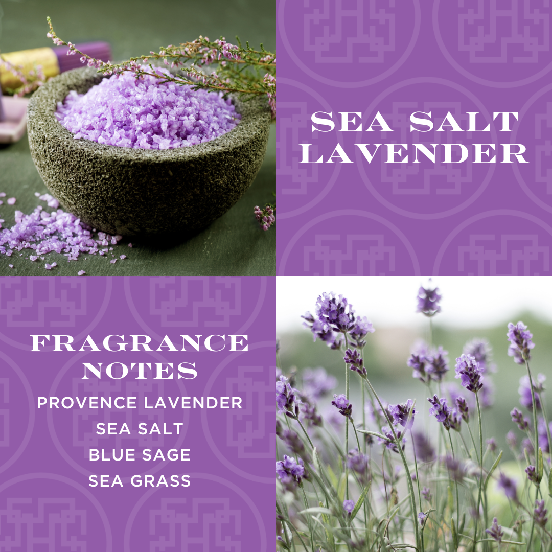 A candle with lavender-colored wax, adorned with a label reading 'SEA SALT LAVENDER', placed on a bed of dried lavender with a small wooden bowl filled with purple crystals next to it. In the background is a field of lavender plants.