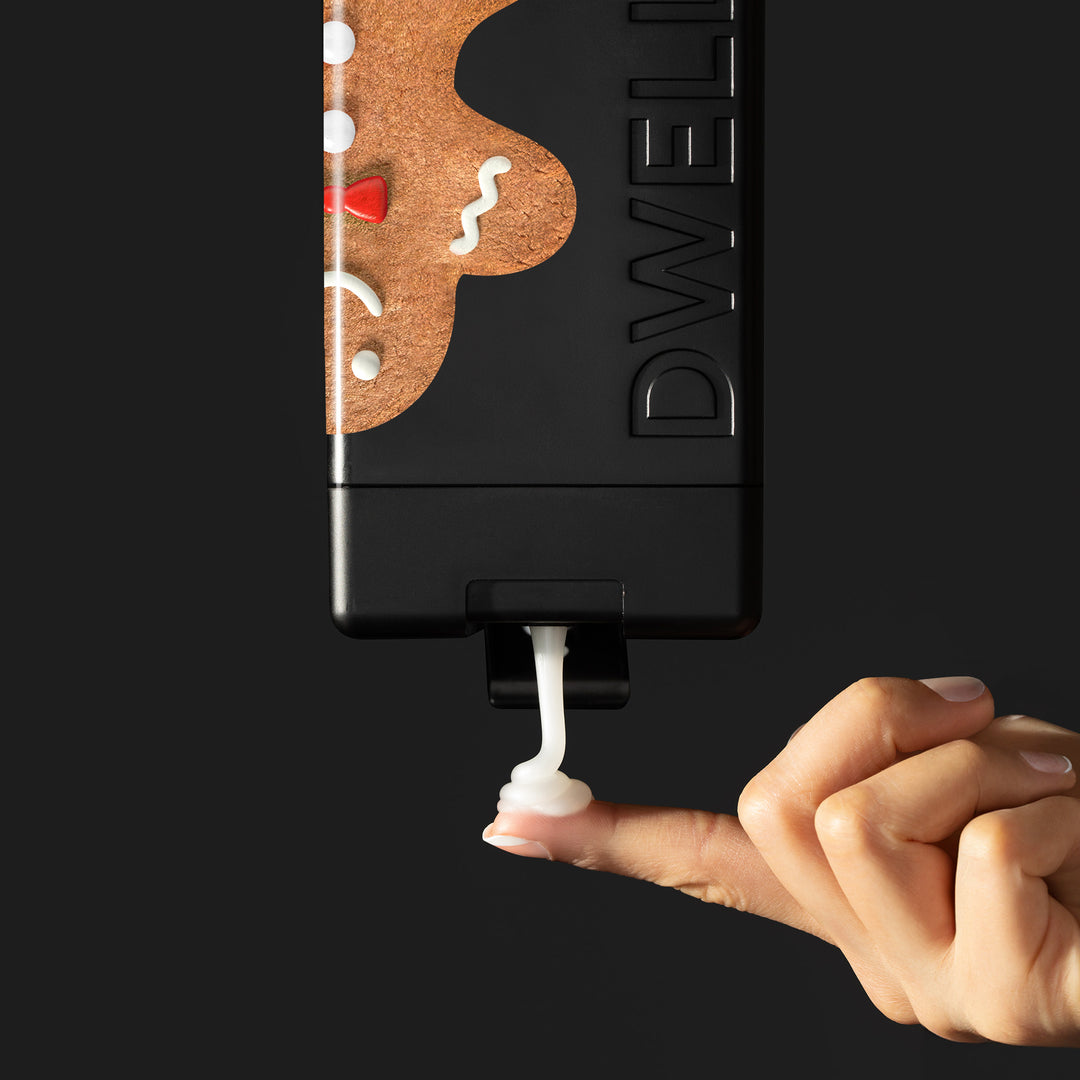 Dwell212 GINGERBREAD body wash bottle with gingerbread man design flipped upside down, being squeeze to dispense product on to the fingertip of an outreached hand