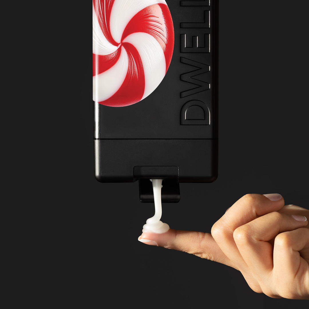 Dwell212 PEPPERMINT body wash bottle with peppermint swirl candy design flipped upside down, being squeeze to dispense product on to the fingertip of an outreached hand
