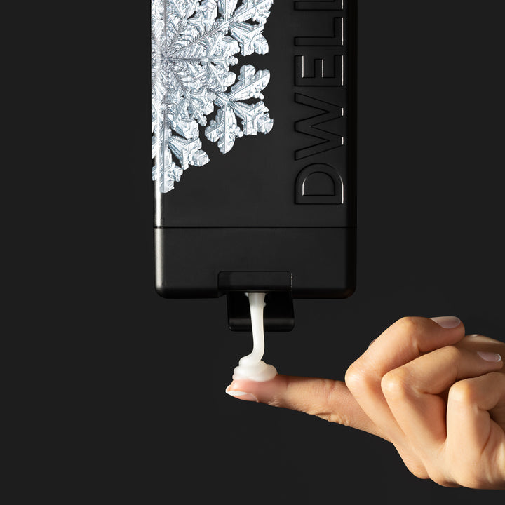 Dwell212 SNOW body wash bottle with snowflake design flipped upside down, being squeeze to dispense product on to the fingertip of an outreached hand